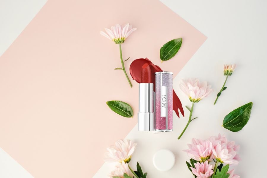 Thỏi son đỏ và hoa cúc hồng trên nền giấy màu hồng nhạt, tạo cảm giác tươi mới và nữ tính.
