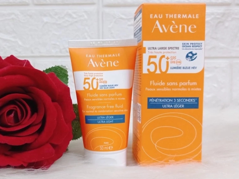 db2445b1-kem-chong-nang-avene-cho-da-kho-2 Tuýp và hộp kem chống nắng đặt trên nền trắng, bên cạnh là bông hoa hồng đỏ.
