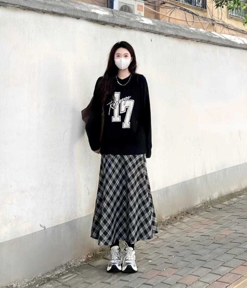 Người đeo khẩu trang, mặc áo thun đen số 17 và váy caro, đứng trước bức tường trắng trên vỉa hè.