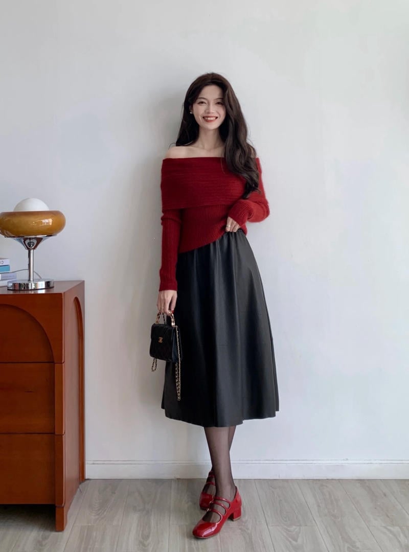 Người phụ nữ mặc áo len đỏ, váy đen, đang đứng dựa vào tường, cầm túi xách nhỏ, cạnh bàn gỗ.