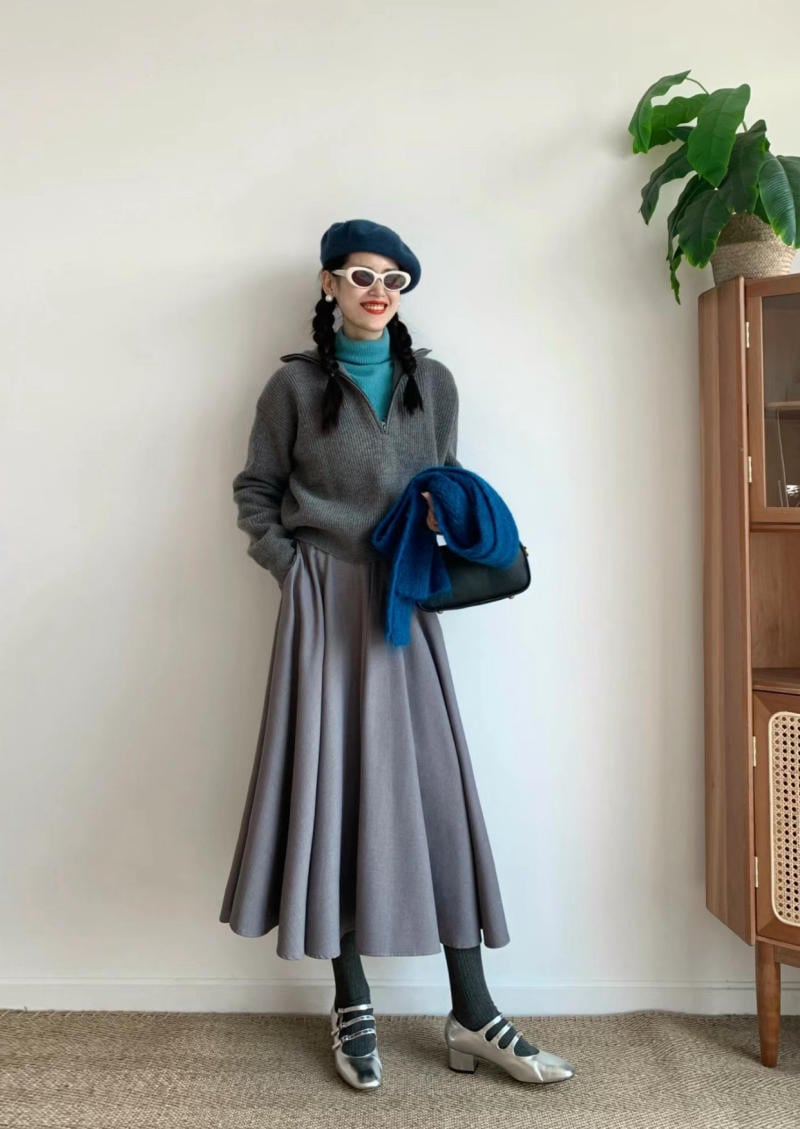 Người phụ nữ mặc áo len xám, váy dài, đội mũ beret xanh, đeo kính trắng, đứng cạnh tủ gỗ và cây xanh.