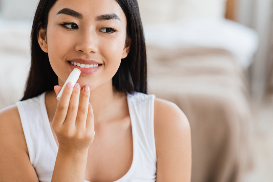 Người phụ nữ đang thoa son dưỡng lên môi, ngồi trong phòng có ánh sáng dịu nhẹ.