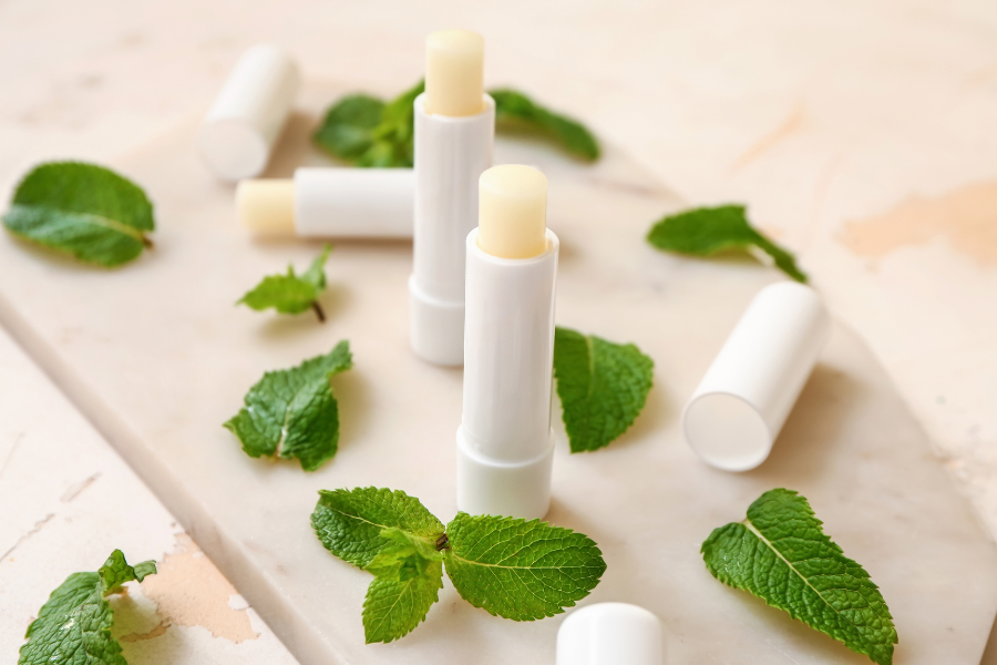 Các thỏi son dưỡng môi trắng đặt trên bề mặt đá cẩm thạch, xung quanh là lá bạc hà tươi.