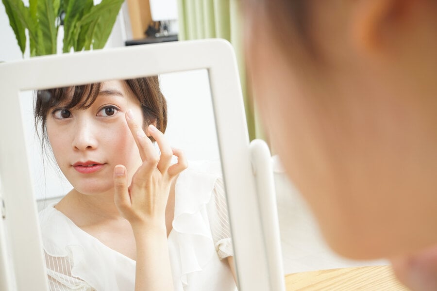 Người phụ nữ đang nhìn vào gương và chạm nhẹ vào vùng da quanh mắt, trong không gian sáng sủa.
