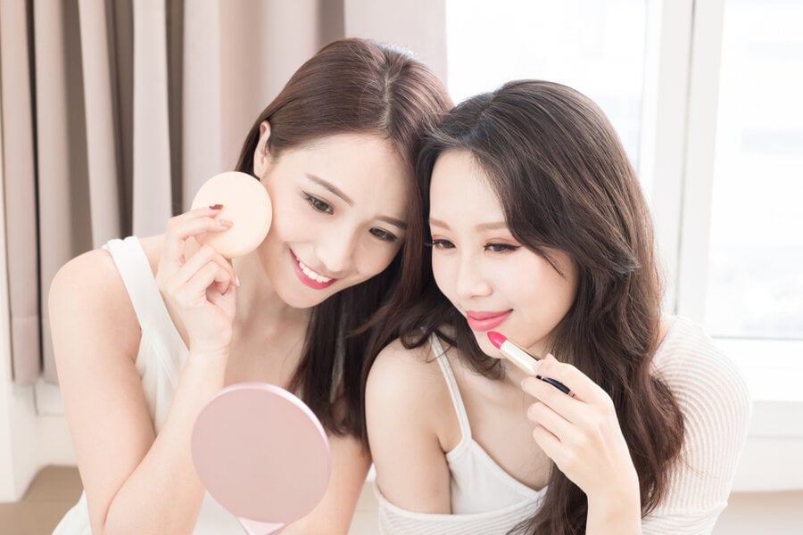 Hai người đang trang điểm trước gương trong phòng sáng sủa, một người dùng cọ phấn, người kia thoa son môi.