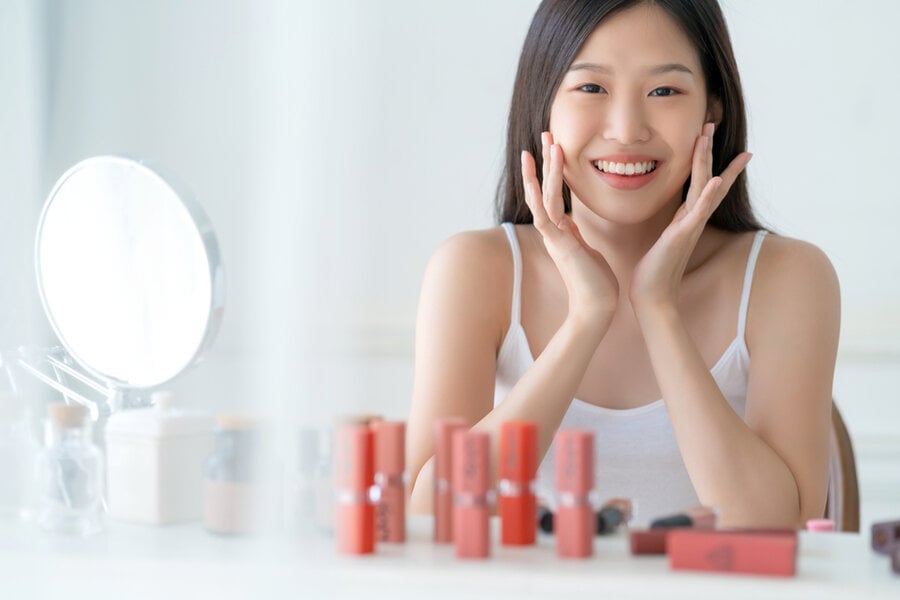 Người phụ nữ mỉm cười ngồi trước gương, xung quanh có nhiều sản phẩm trang điểm trên bàn.