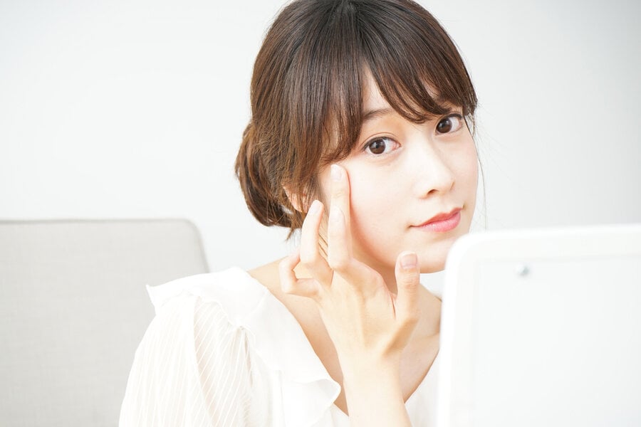 Người phụ nữ chạm vào mặt khi nhìn vào gương, ngồi trên ghế với áo trắng trong nền sáng.