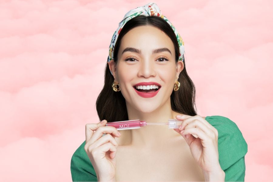 Người phụ nữ cầm son bóng hồng, mặc áo xanh, nền màu hồng, cười tươi.