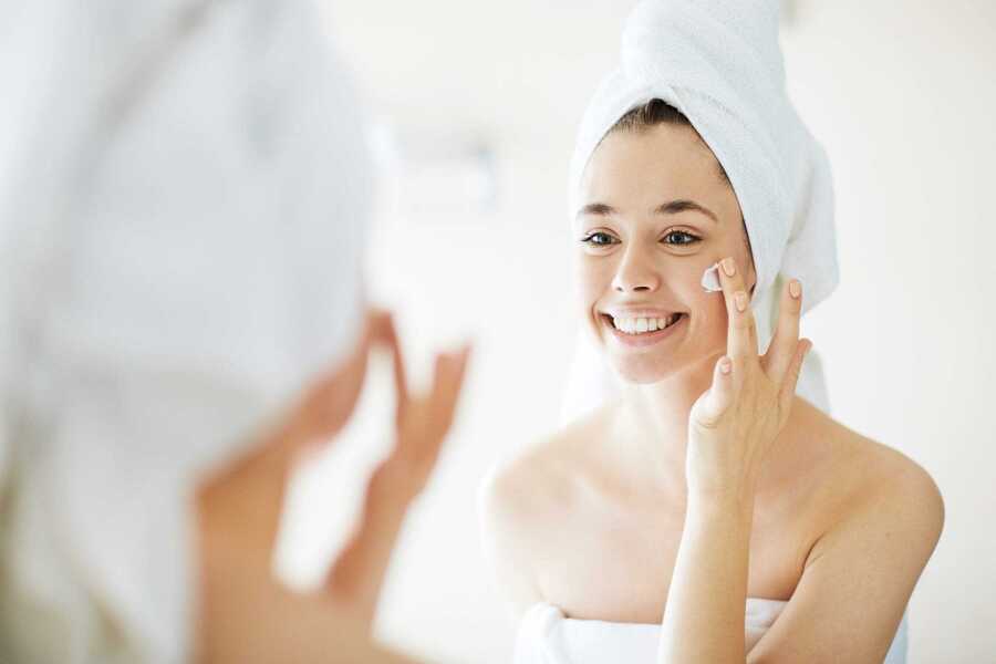 Người phụ nữ quấn khăn tắm, thoa kem lên mặt trước gương, biểu cảm tươi cười, không gian sáng sủa.