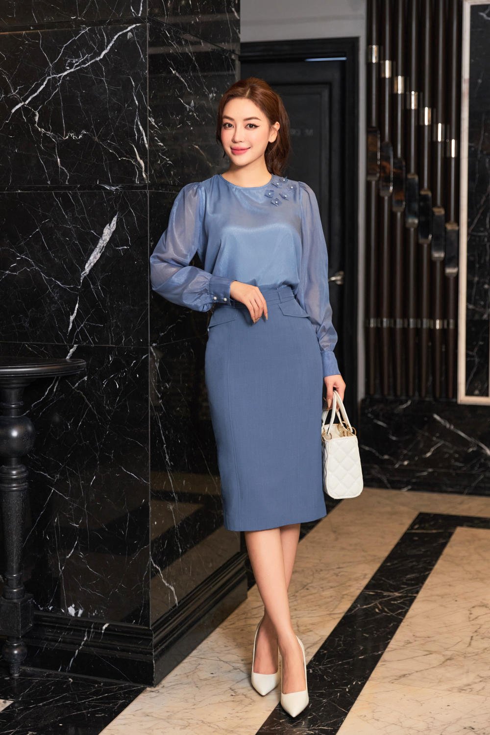 Người phụ nữ mặc trang phục xanh đứng dựa vào tường đá đen, tay cầm túi xách trắng trong không gian sang trọng.