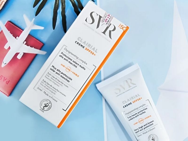 Hộp và tuýp kem chống nắng SPF50+ đặt trên nền xanh, bên cạnh mô hình máy bay và hộ chiếu đỏ.