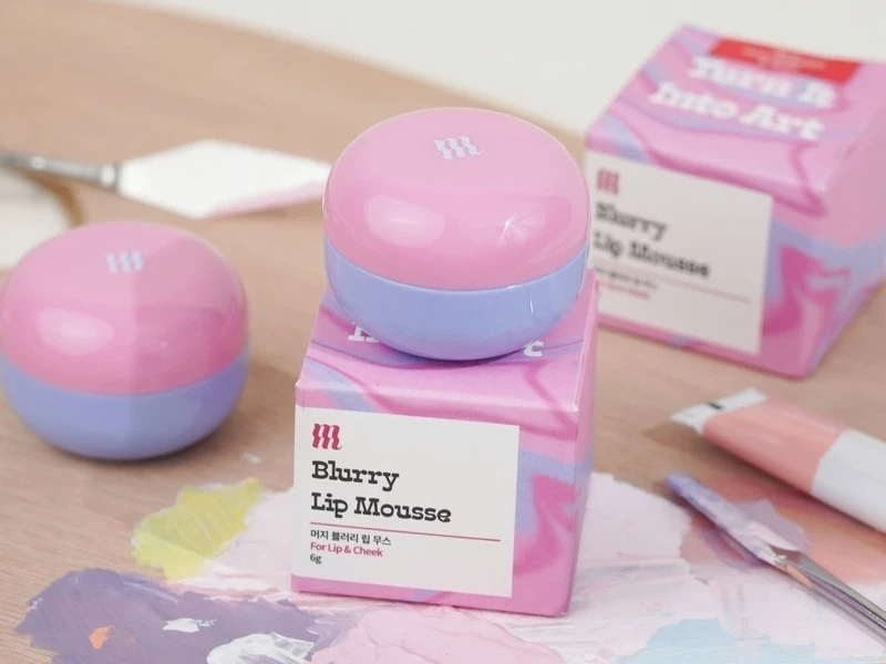 e8e869f7-son-merzy-mau-nao-dep-nhat-27 Hộp và hũ kem màu hồng và tím nhạt trên bàn gỗ, kèm dụng cụ vẽ và màu sắc trộn lẫn.