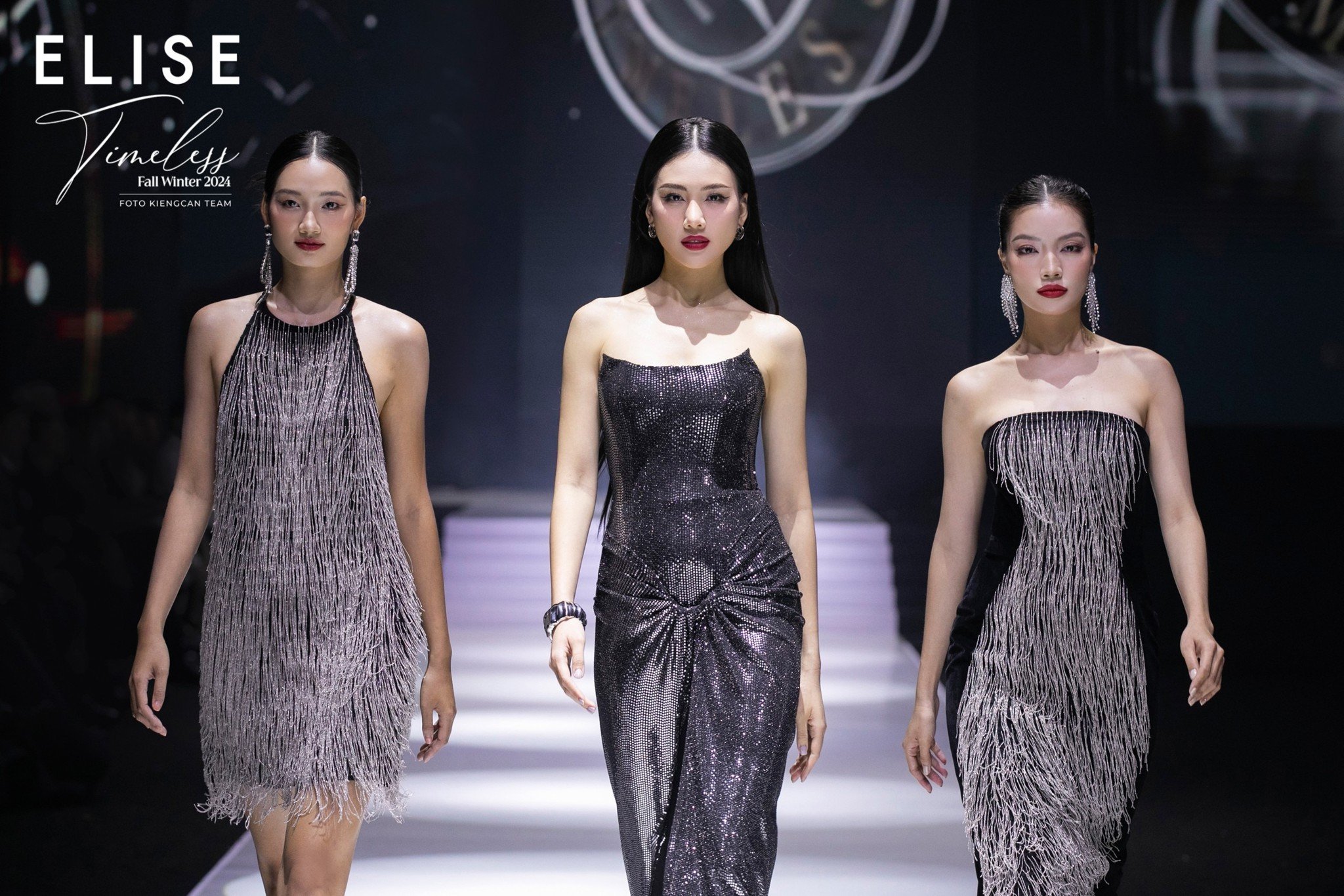 Ba người mẫu trình diễn trang phục dạ hội lấp lánh trên sàn diễn thời trang, nền tối và ánh sáng nổi bật.