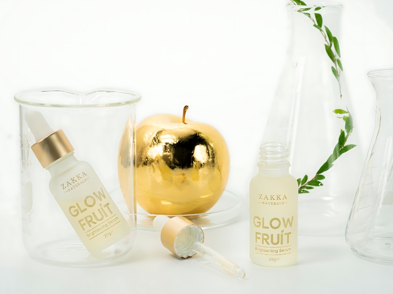 Chai serum và ống nhỏ giọt bên cạnh quả táo mạ vàng và cốc thủy tinh trong suốt, nền trắng.