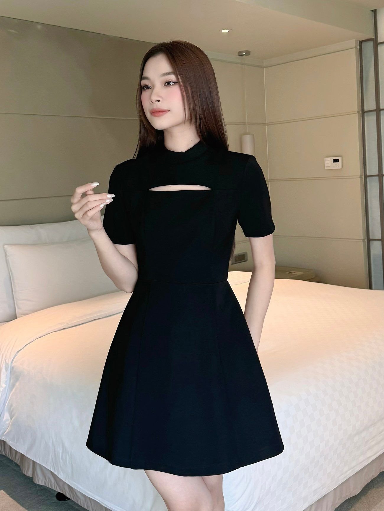 Người phụ nữ mặc váy đen đứng trong phòng với giường trắng phía sau.