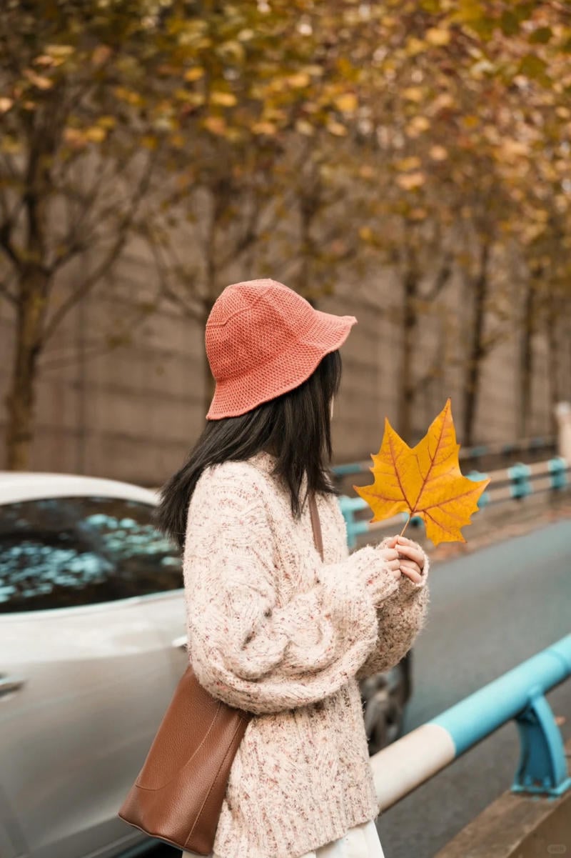 Người mặc áo len và mũ, cầm lá vàng, đứng cạnh xe hơi trên đường có hàng cây mùa thu.