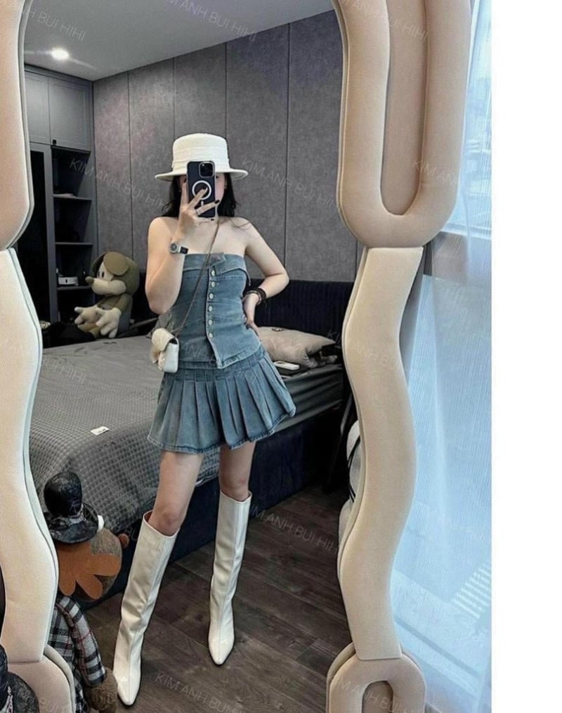 Người chụp selfie trước gương, mặc váy denim, đội mũ trắng và đi bốt cao, trong phòng ngủ hiện đại.