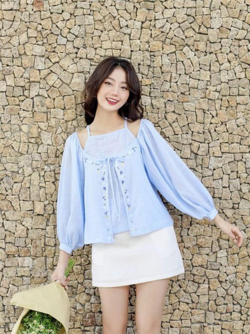 Người phụ nữ mặc áo xanh nhạt và váy trắng, đứng trước bức tường đá, cầm bó hoa trong tay.