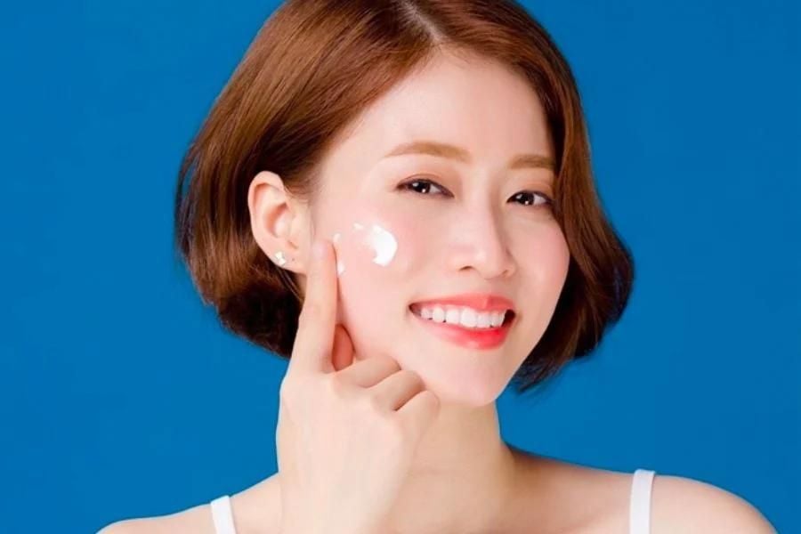 Người phụ nữ đang mỉm cười, thoa kem dưỡng da lên má, nền màu xanh dương, mái tóc ngắn.