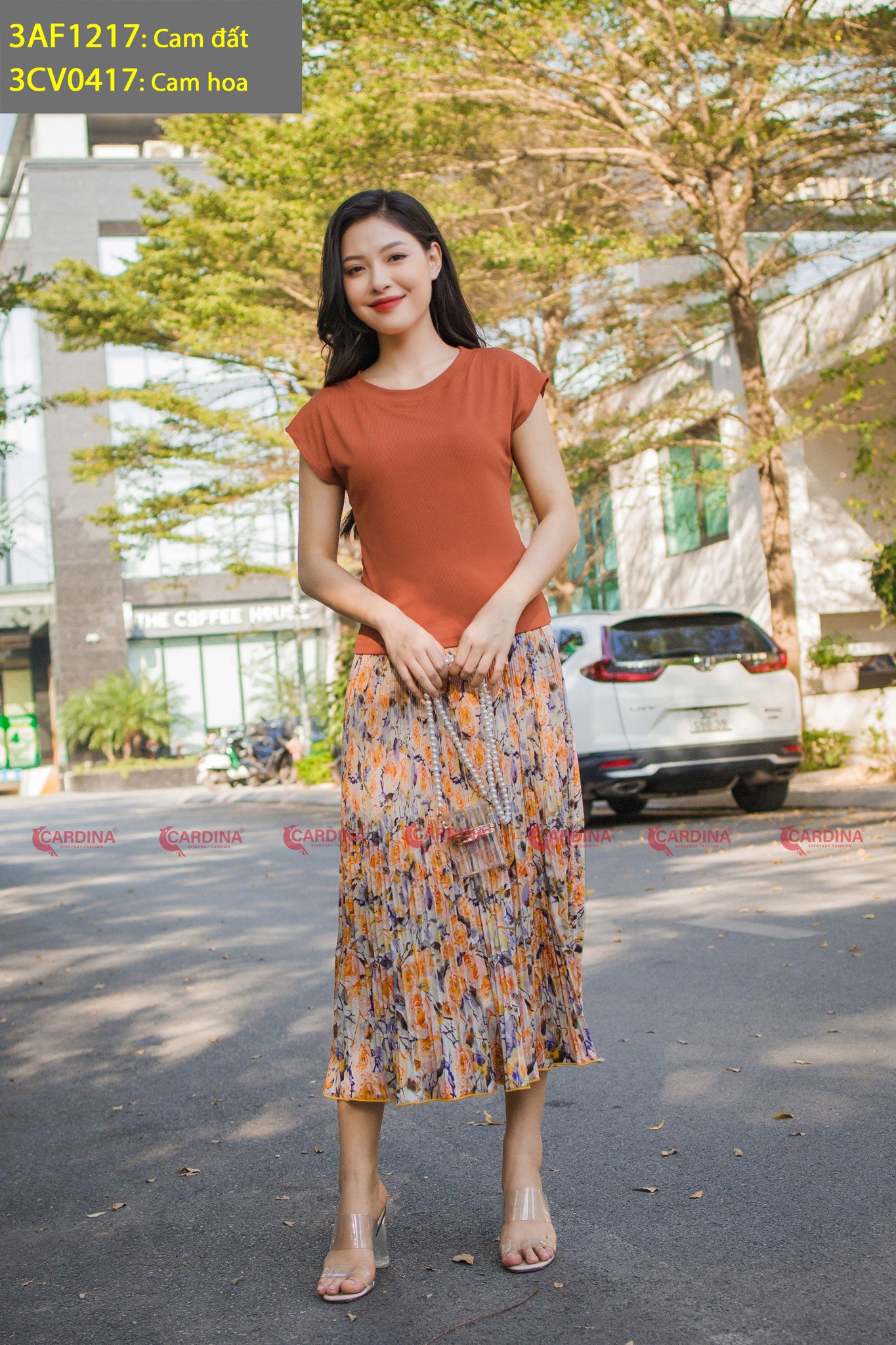 10 Cách Phối Đồ Với Chân Váy Hoa Nhí Giúp Bạn Tỏa Sáng Mùa Hè 10 Người phụ nữ mặc áo cam và váy hoa đứng trên đường, phía sau là cây cối và ô tô.
