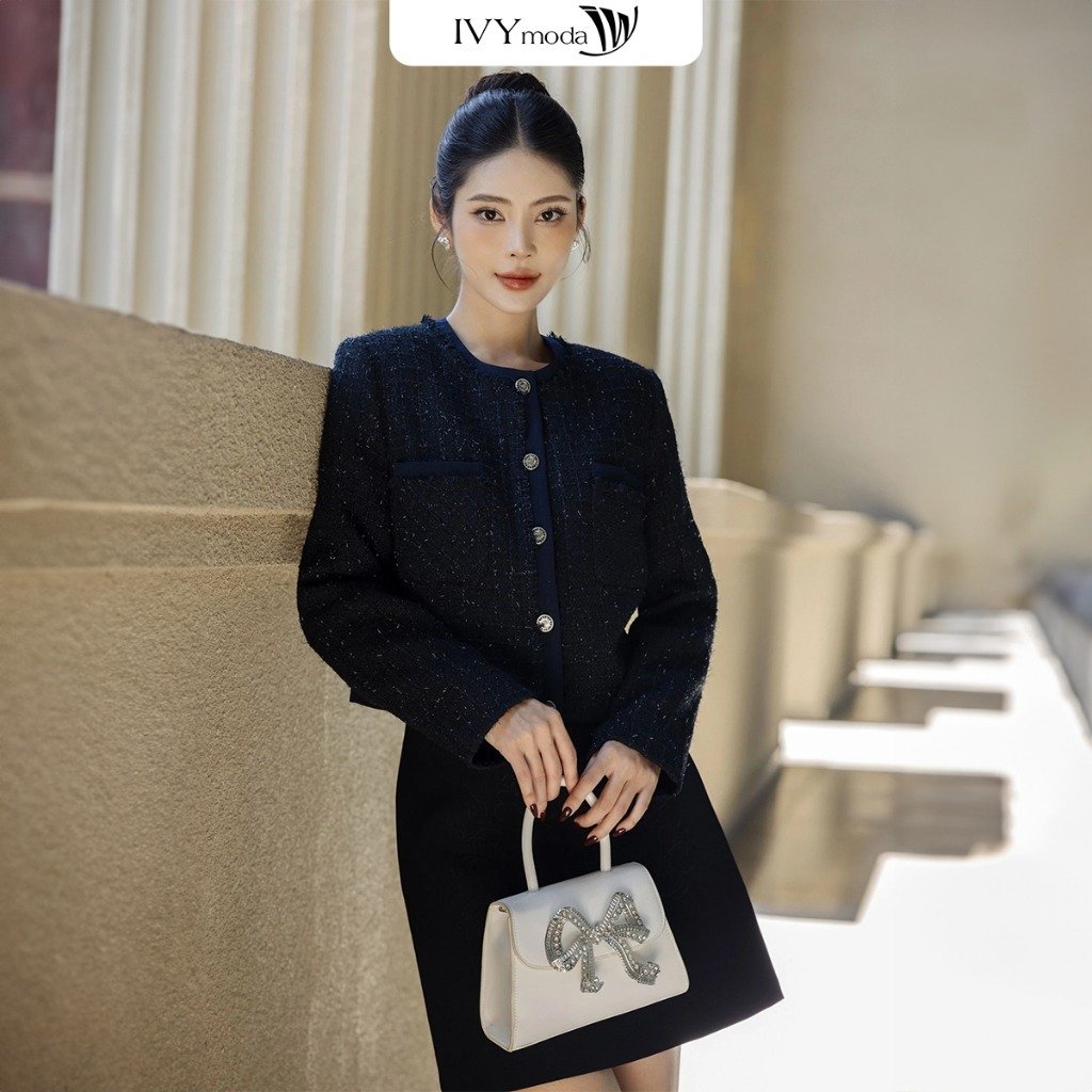 Người phụ nữ mặc trang phục đen, cầm túi xách trắng đứng dựa vào bức tường, phía sau là cột trụ lớn.