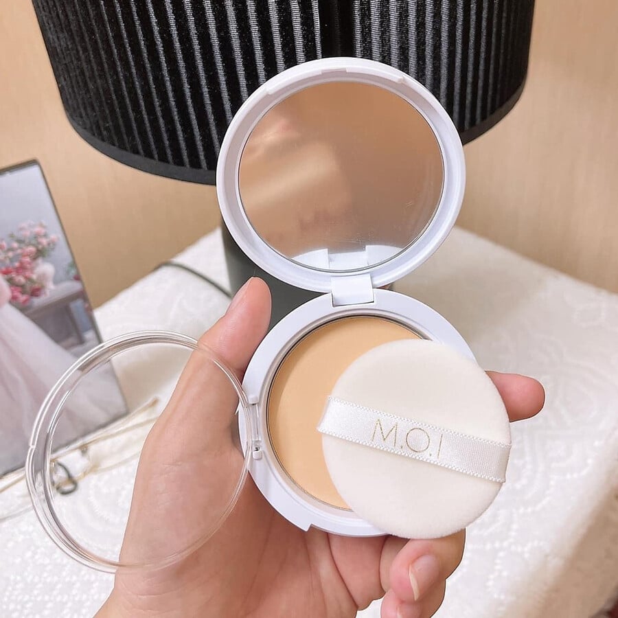 Hộp phấn phủ với bông phấn được cầm trên tay, đặt trên nền trắng với đèn trang trí phía sau.