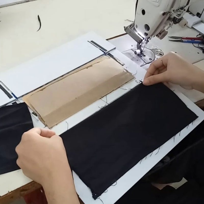 Người đang điều chỉnh vải đen trên máy may, bên cạnh là hộp các tông và một số dụng cụ.