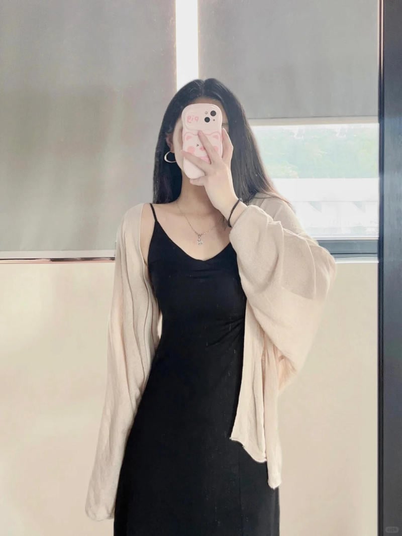 Người đang tự chụp ảnh trước gương, mặc váy đen và áo khoác màu sáng, che mặt bằng điện thoại.