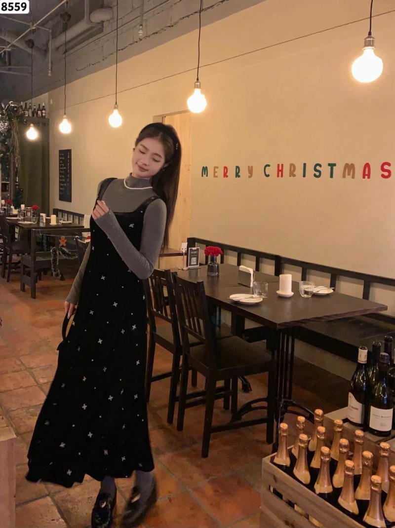 Người phụ nữ đứng trong nhà hàng trang trí Giáng Sinh, mặc váy dài đen, bên cạnh là bàn ăn và đèn sáng.