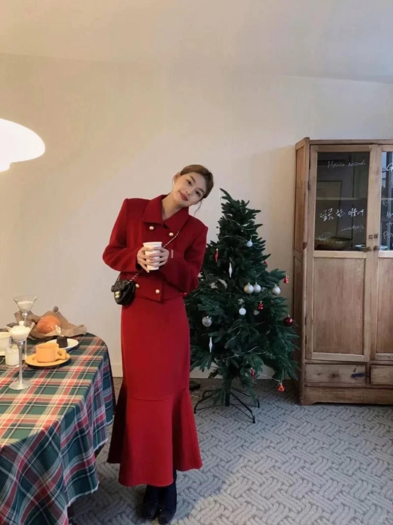 Người phụ nữ mặc trang phục đỏ, cầm cốc, đứng cạnh cây thông Noel và bàn có khăn trải kẻ sọc.