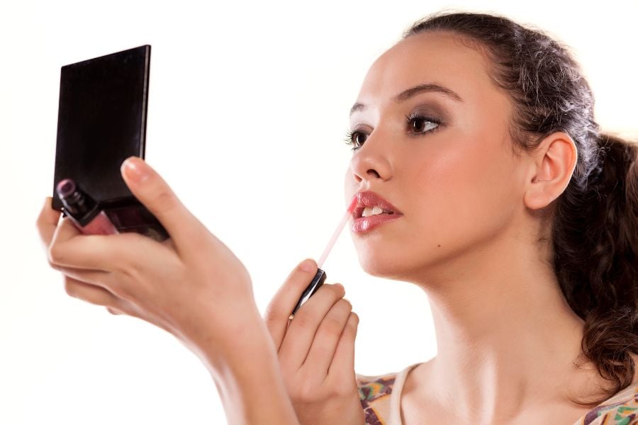 Người phụ nữ đang thoa son môi, tay cầm gương nhỏ, tập trung nhìn vào gương để trang điểm.
