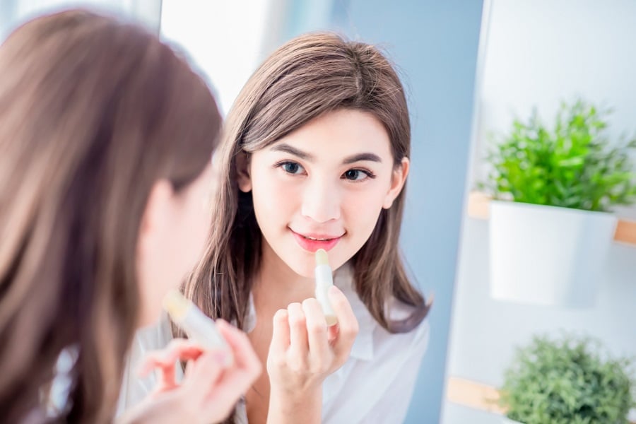 Người phụ nữ đang soi gương và thoa son dưỡng môi, phía sau là cây xanh trang trí.