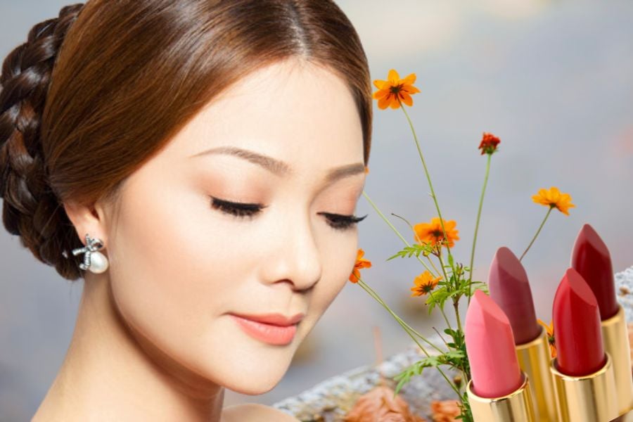 Người phụ nữ trang điểm nhẹ nhàng, tóc búi, bên cạnh có hoa và ba thỏi son màu đỏ, hồng.