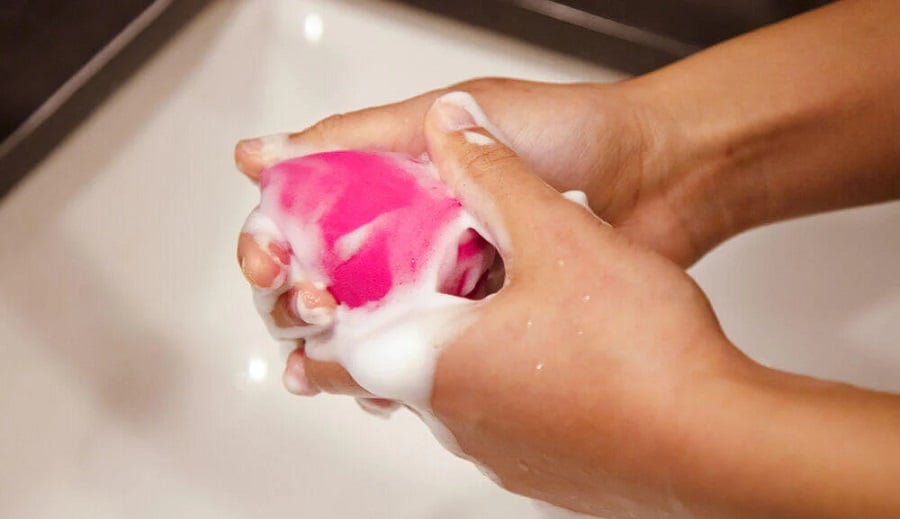 Hai tay đang rửa một miếng bọt biển trang điểm màu hồng với xà phòng trong bồn rửa.