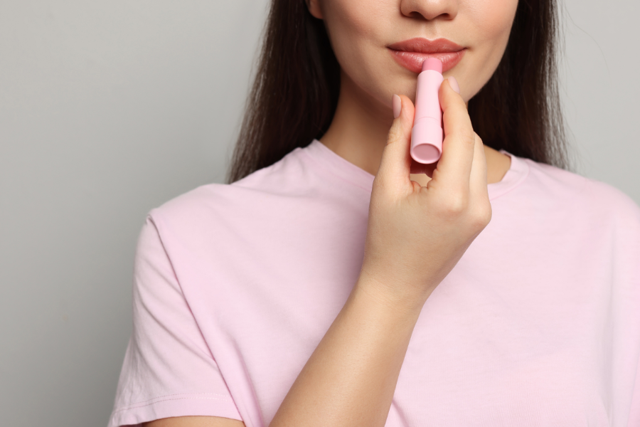 Người mặc áo hồng đang thoa son dưỡng môi màu hồng nhạt lên môi, nền xám nhạt.