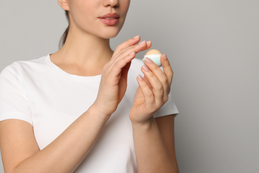 Người mặc áo trắng cầm và mở nắp một hộp sáp dưỡng môi màu xanh nhạt trên nền xám.