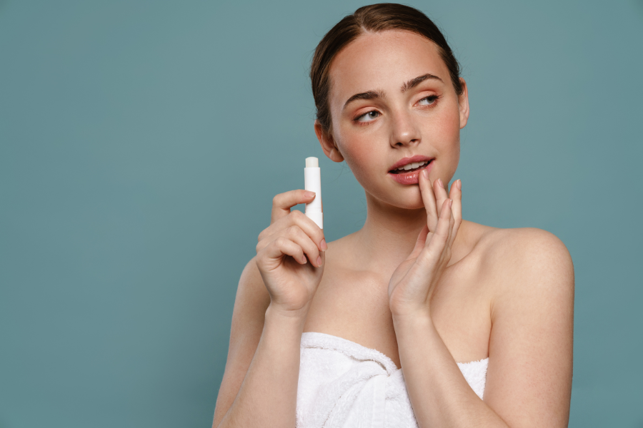 Người cầm sản phẩm chăm sóc môi, quấn khăn trắng, nền xanh dương, biểu cảm suy tư.