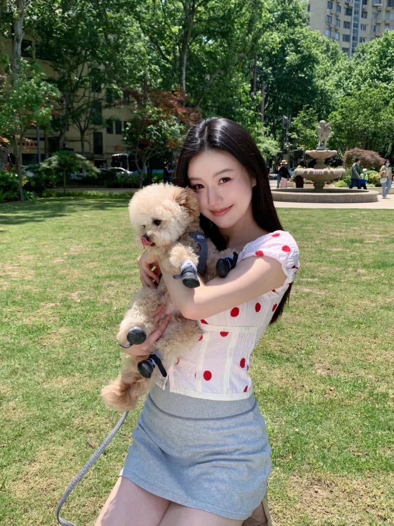 Người phụ nữ ôm chú chó nhỏ trong công viên xanh, phía sau là đài phun nước và các tòa nhà.