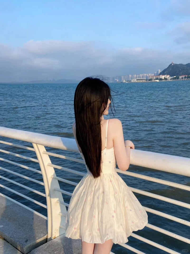 Người đứng nhìn ra biển từ lan can, mặc váy trắng, trời trong xanh và có dãy nhà phía xa.