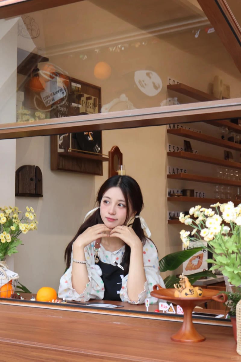 Người phụ nữ ngồi bên cửa sổ quán cà phê, xung quanh có hoa và đồ trang trí.