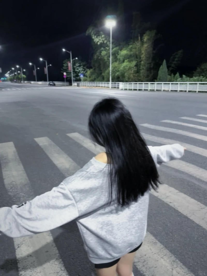Người đang dang tay đứng trên vạch qua đường vào ban đêm, với đèn đường sáng và cây xanh phía xa.