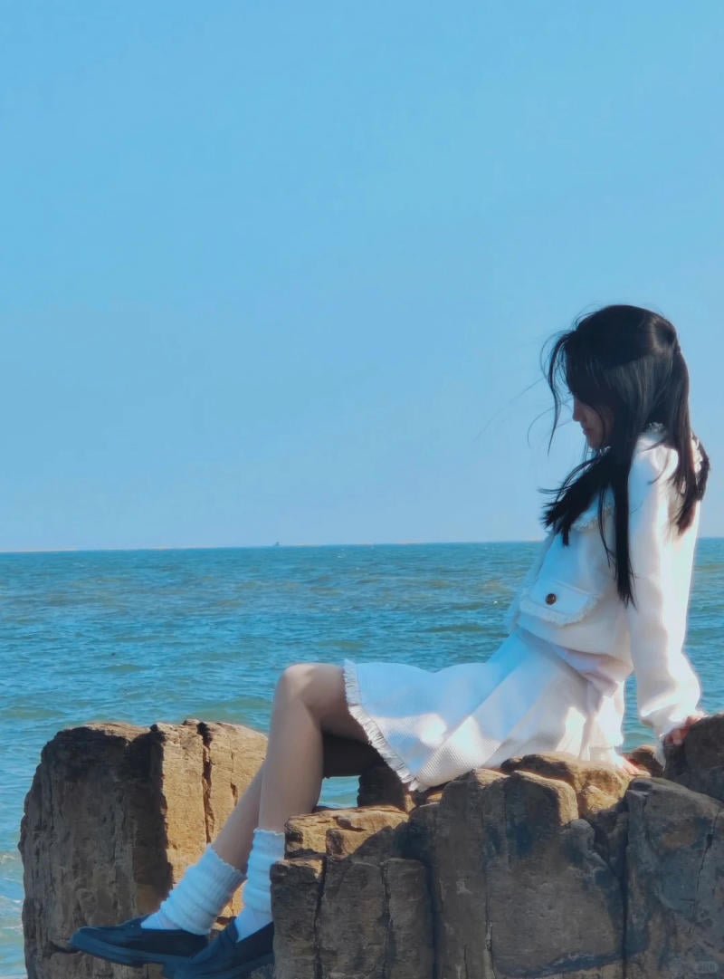 Người ngồi trên tảng đá gần biển, mặc trang phục trắng, nhìn xa xăm về phía đường chân trời.
