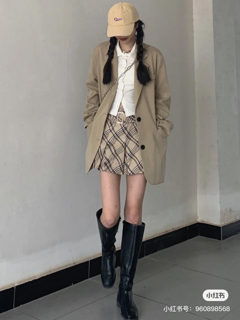 Người mẫu mặc áo khoác be, váy kẻ caro, đội mũ lưỡi trai, đi bốt đen, đứng dựa vào tường trong nhà.
