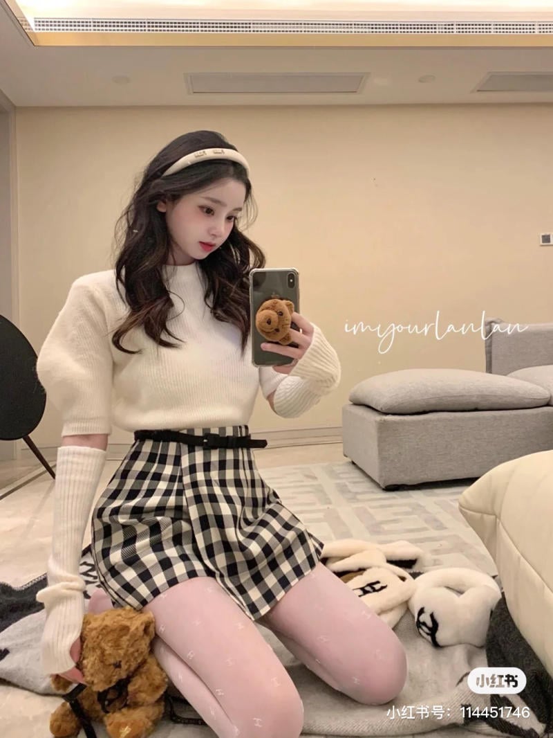 Người chụp ảnh selfie trong phòng, mặc áo trắng và váy caro, tay cầm điện thoại có ốp hình gấu.