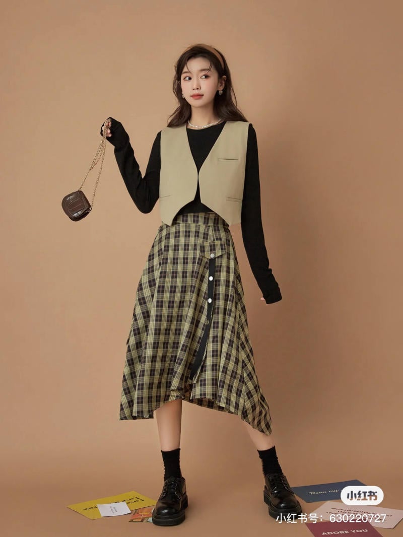 Người mẫu mặc áo gile, áo dài tay và váy kẻ caro, cầm túi nhỏ, đứng trên nền màu nâu.