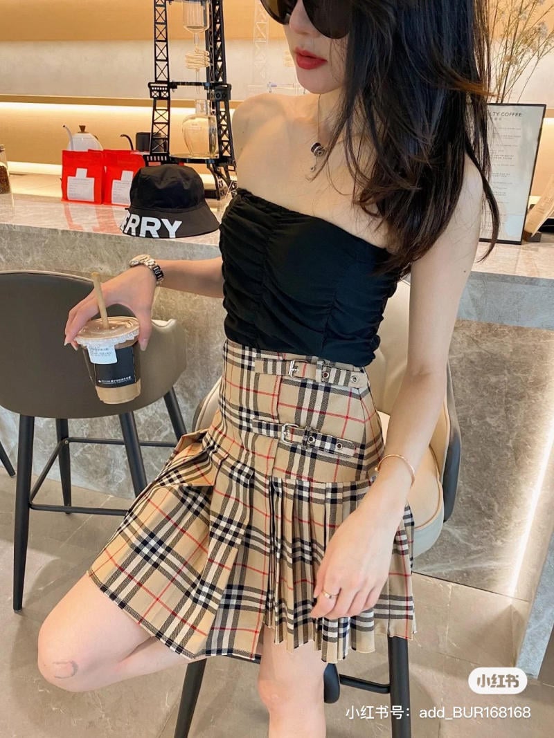 Người phụ nữ mặc áo đen không tay và váy kẻ caro, cầm ly nước, đứng cạnh quầy bar.