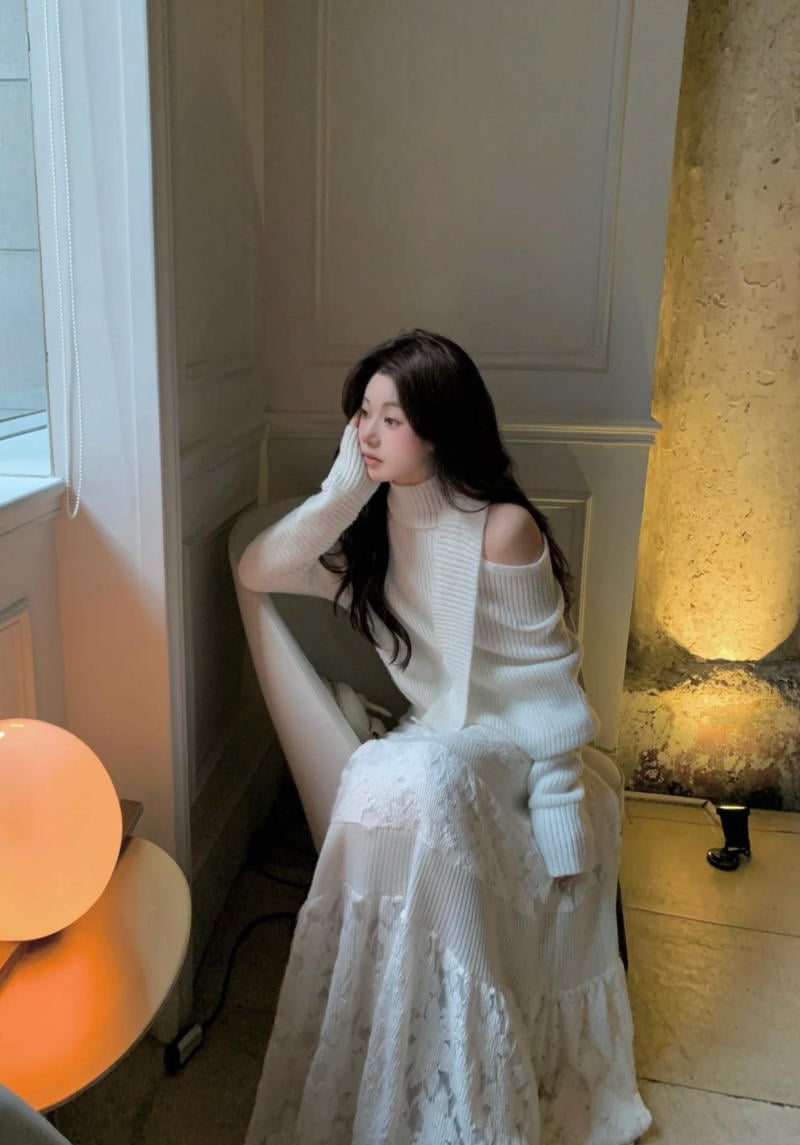 Người ngồi trên ghế trong phòng, mặc áo len trắng, nhìn ra cửa sổ với ánh sáng dịu nhẹ từ đèn bàn bên cạnh.