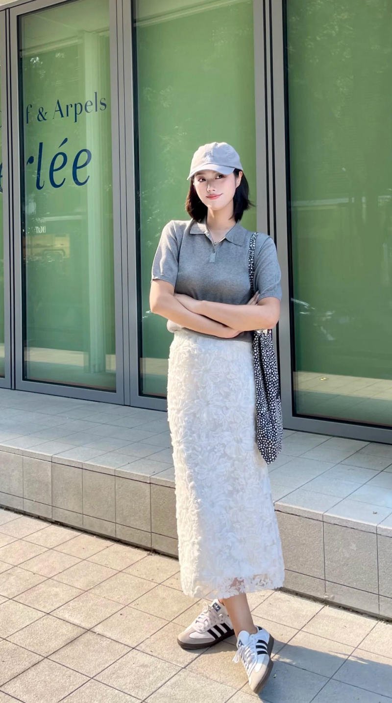 Người phụ nữ đội mũ lưỡi trai, áo xám, váy trắng đứng trước cửa kính, tay cầm túi vải chấm bi.
