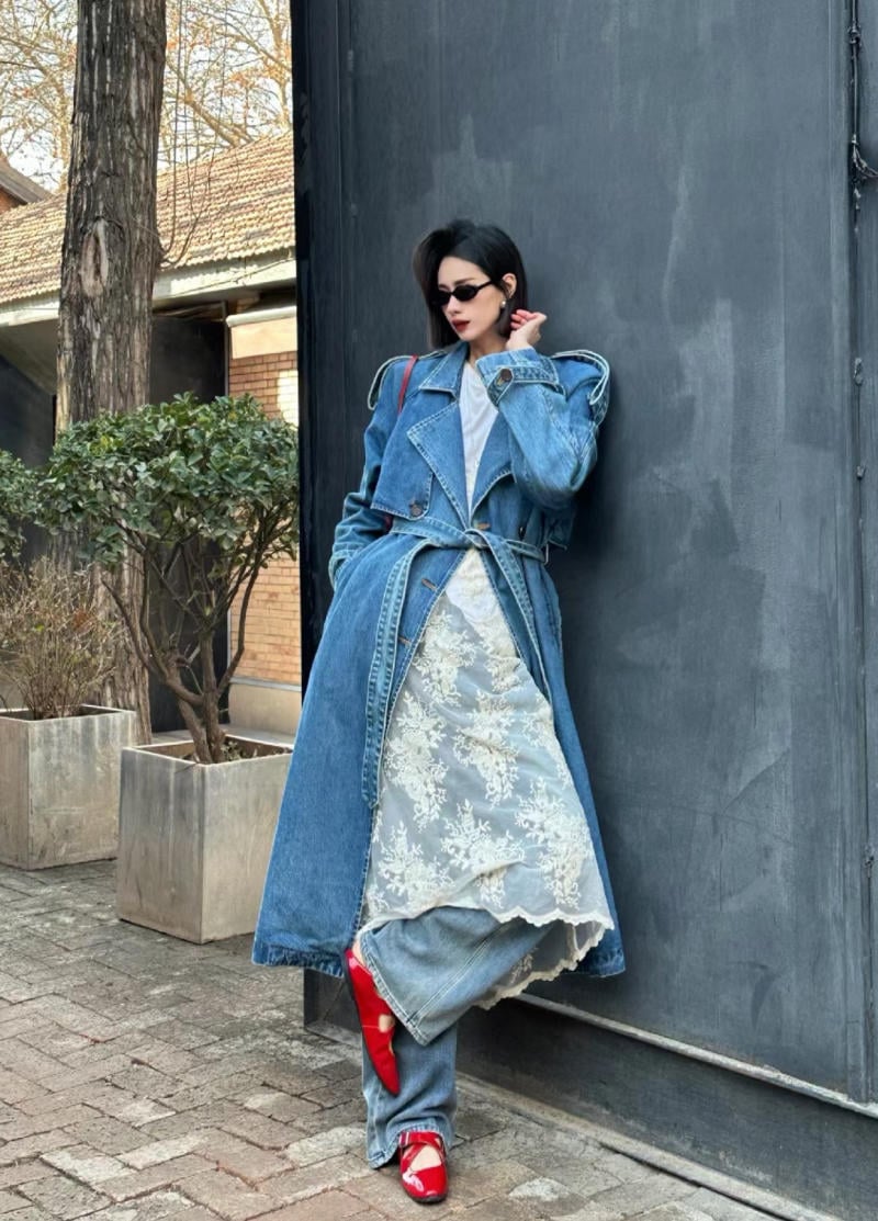 Người mặc áo khoác denim dài, đầm ren trắng và giày đỏ, đứng dựa lưng vào tường ngoài trời.