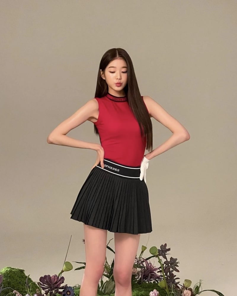 Người mẫu mặc áo đỏ và váy đen đứng tạo dáng, xung quanh là hoa và cây cảnh.